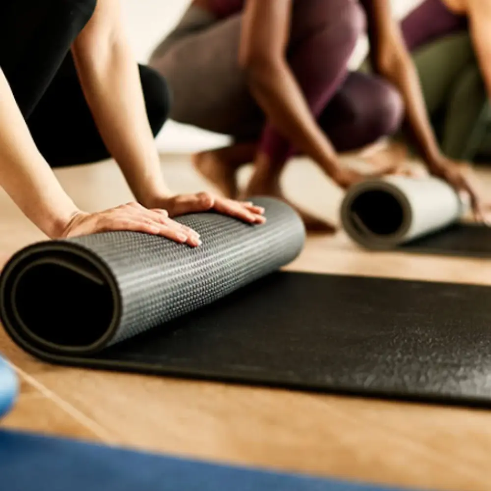 A group of people rolling up black and gray Pilates Mats on a wooden studio floor.